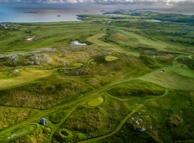 A scenic golf course with lush green fairways, surrounded by rugged hills and adjacent to a calm blue ocean under a partly cloudy sky.