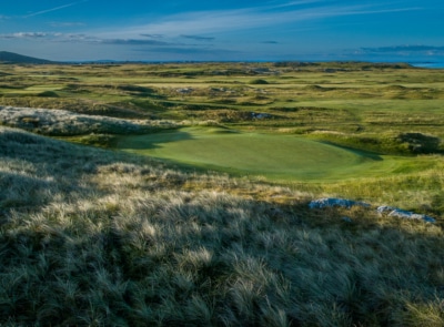 A picturesque golf course nestled along the rugged coastline, featuring rolling green fairways, sandy bunkers, and the shimmering ocean under a clear blue sky.