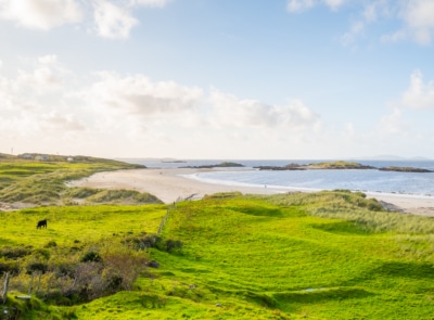A serene sandy beach with gentle waves, surrounded by rocky hills and lush greenery under a clear blue sky in Connemara, Co. Galway.