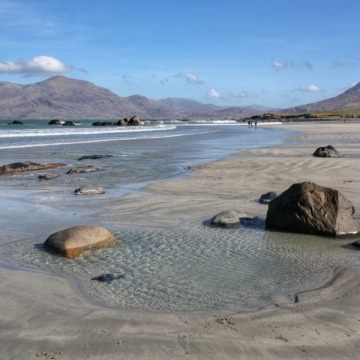 A scenic beach with golden sand, rocky shoreline, and turquoise water beneath a cloudy sky, surrounded by lush greenery in Connemara, County Galway.