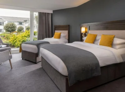 A bright, elegantly furnished hotel room with twin beds featuring white linens, a seating area, and large windows overlooking a lush garden with trees and outdoor seating.