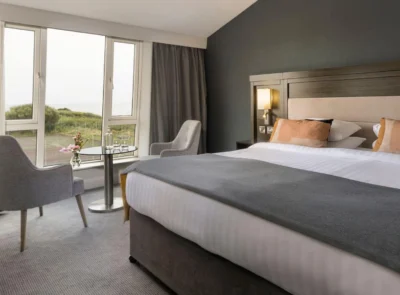 A luxurious hotel room with a large bed, elegant decor, and a scenic ocean view through a wide window, featuring soothing, neutral colour tones.