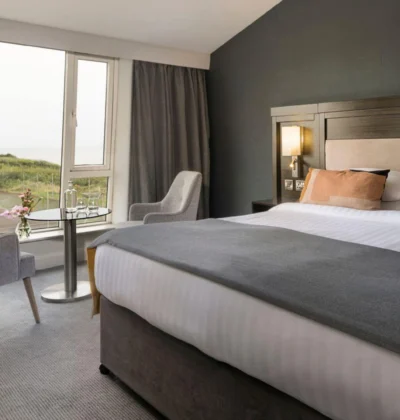 A luxurious hotel room with a large bed, elegant decor, and a scenic ocean view through a wide window, featuring soothing, neutral colour tones.