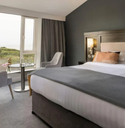 A luxurious hotel room with a large bed, elegant decor, and a scenic ocean view through a wide window, featuring soothing, neutral colour tones.