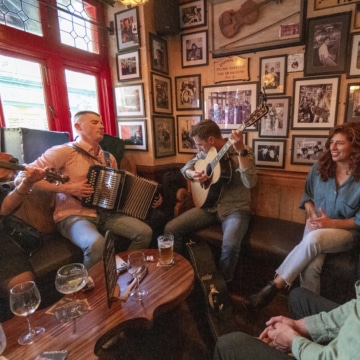 Image of a lively Irish pub with patrons enjoying traditional music. The pub has wooden interiors, adorned with various memorabilia, and musicians are seen performing in the background.