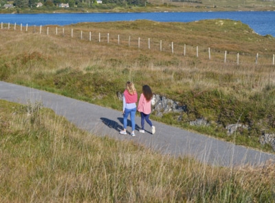 A scenic view of a lush green pathway lined with trees and vegetation, under a clear sky, inviting walkers to explore the tranquil nature of Connemara Greenway, Galway.