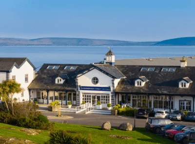 A beautiful coastal hotel with modern architecture surrounded by lush greenery, overlooking the serene ocean waters with clouds scattered across the sky.