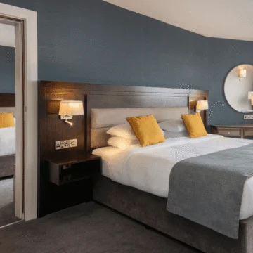 A hotel room with two beds, stylish decor, carpeted floor, bedside lamps, and large windows offering a scenic view over a body of water.