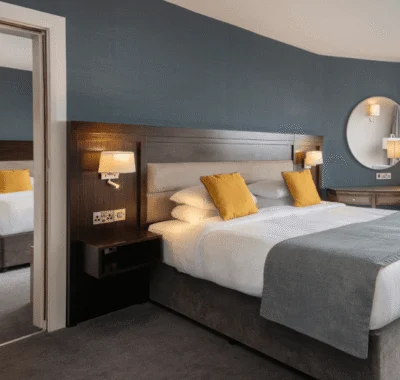 A hotel room with two beds, stylish decor, carpeted floor, bedside lamps, and large windows offering a scenic view over a body of water.