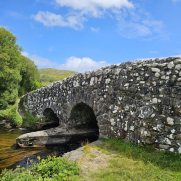 Stone bridge with three arches spans over a calm river in a lush, green landscape under a cloudy blue sky in Connemara, County Galway, Ireland.