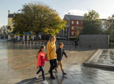 A lively street scene in Galway City with colorful shops, pedestrians strolling, and people dining outdoors under blue skies and fluffy white clouds.