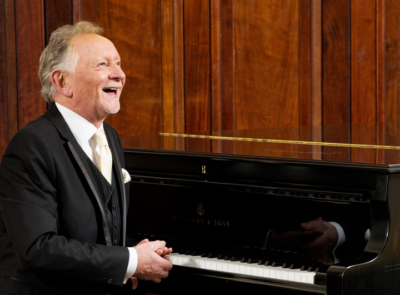 A serene coastal landscape with a calm sea, rocky shoreline, and a clear blue sky, possibly associated with Phil Coulter, likely promoting an event or location.