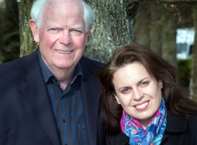 A man and a woman, both smiling. The man, elderly with white hair, wears a dark suit. The woman has long dark hair and wears a red dress.