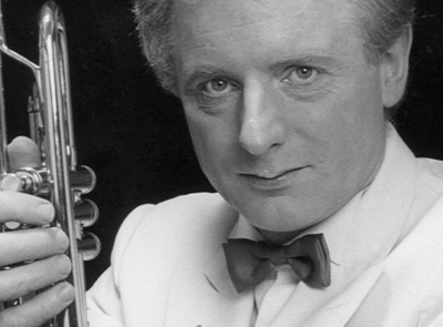 A man playing a trumpet, wearing a black outfit, with a blurred green background, possibly outdoors. His eyes are focused on the instrument, emphasizing his musical engagement.