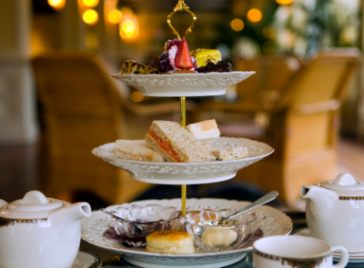 Afternoon tea setup with a variety of pastries, sandwiches, scones, and tea served on elegant crockery in a bright room with a scenic window view.