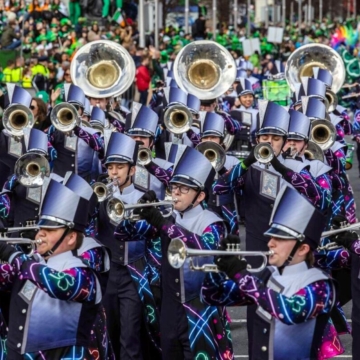 A serene coastal landscape with rocky shoreline, calm water, and distant mountains under a clear blue sky. Green grass and wildflowers in the foreground enhance the peaceful setting.