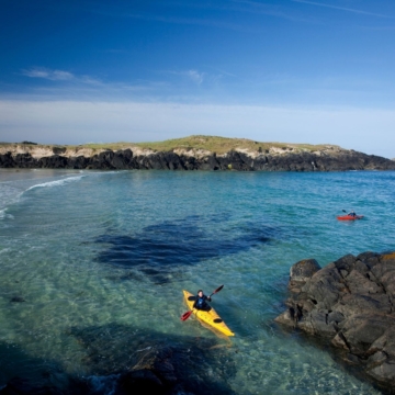 Aerial view of Connemara Coast Hotel surrounded by green landscapes, bordered by a rocky shoreline, and overlooking a calm blue sea under a clear sky.