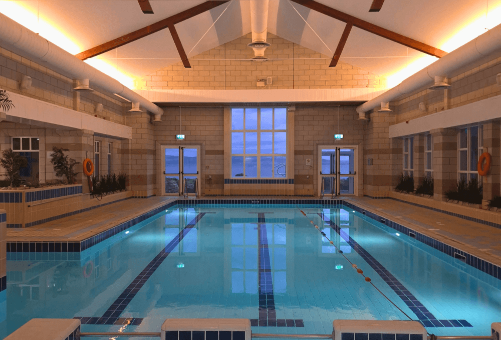 Guests enjoying serene indoor pool with skylight, warm lighting, and sea views. Relaxing swim experience at seaside hotel.