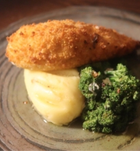 A plate with a golden-brown chicken Kiev served alongside steamed vegetables and a mound of mashed potatoes, garnished with fresh herbs.
