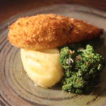 A plate with a golden-brown chicken Kiev served alongside steamed vegetables and a mound of mashed potatoes, garnished with fresh herbs.
