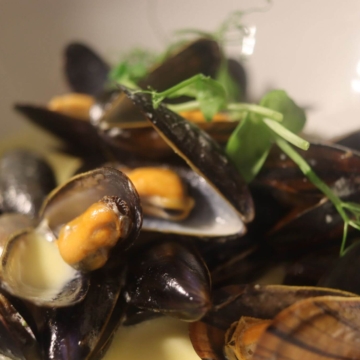 A plate of cooked mussels served in their shells, garnished with herbs and lemon slices, positioned on an elegant white tablecloth.