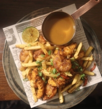 A takeout box filled with a spice bag: fried chicken pieces, sliced chili peppers, onions, and vibrant peppers, displayed on a wooden table.
