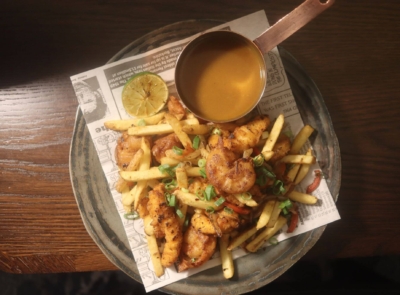 A takeout box filled with a spice bag: fried chicken pieces, sliced chili peppers, onions, and vibrant peppers, displayed on a wooden table.