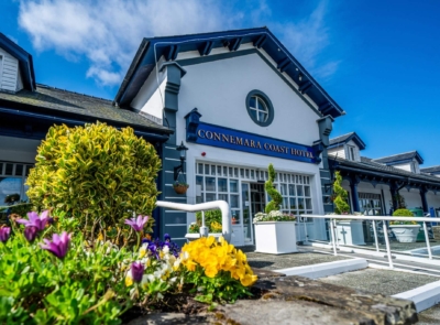 Entrance of Connemara Coast Hotel with a glass-covered walkway, surrounded by well-maintained greenery and coastal views under a clear blue sky.