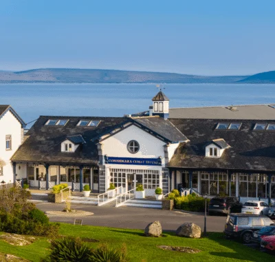 Coastal hotel with a rustic exterior, surrounded by lush greenery and overlooking the sea under a partly cloudy sky. Wooden benches and tables are arranged on the lawn.
