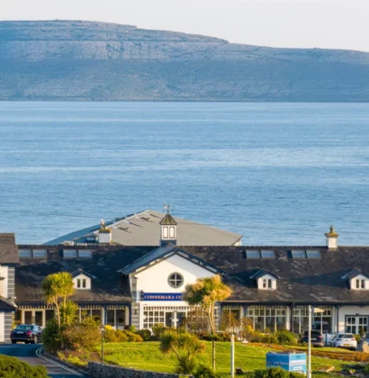 Exterior view of Connemara Coast Hotel, a large white building with a sloping green lawn and vibrant flowers in the foreground under a clear blue sky.