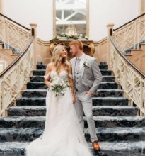 Couple holding hands, walking down stairs with decorative greenery, suited man and woman in white dress, at outdoor wedding venue.