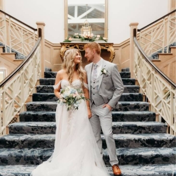Couple holding hands, walking down stairs with decorative greenery, suited man and woman in white dress, at outdoor wedding venue.