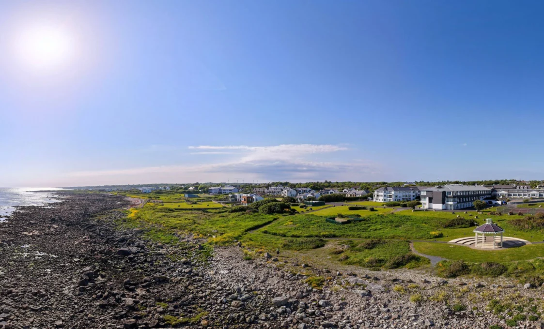 A coastal hotel with large windows, surrounded by trees and overlooking a rocky shoreline and serene sea under a cloudy sky.