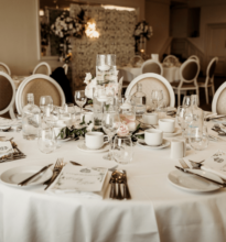 A beautifully decorated wedding reception setup with elegantly arranged tables, white chairs, floral centerpieces, and soft lighting in a spacious event hall.