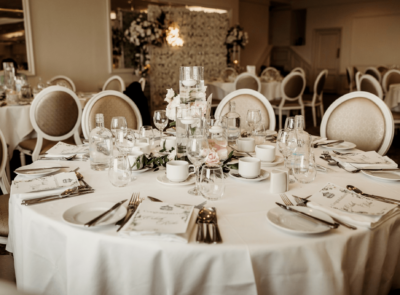 A beautifully decorated wedding reception setup with elegantly arranged tables, white chairs, floral centerpieces, and soft lighting in a spacious event hall.