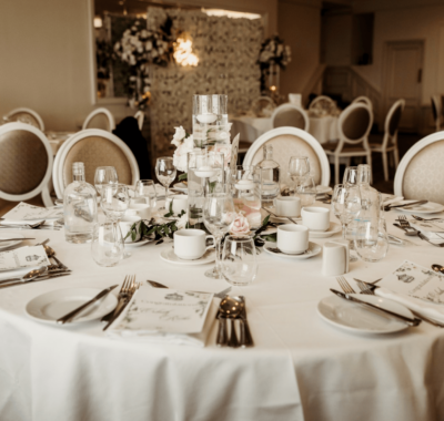 A beautifully decorated wedding reception setup with elegantly arranged tables, white chairs, floral centerpieces, and soft lighting in a spacious event hall.