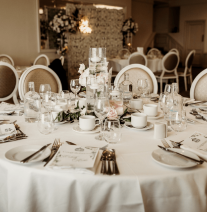 A beautifully decorated wedding reception setup with elegantly arranged tables, white chairs, floral centerpieces, and soft lighting in a spacious event hall.