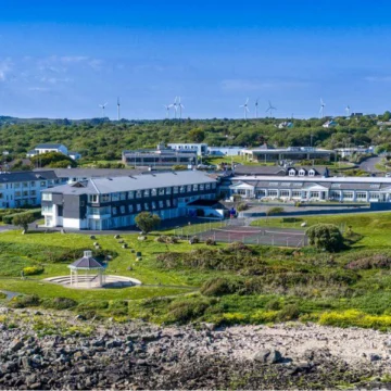 Rear view of Connemara Coast Hotel overlooking scenic coastline, with green lawn, outdoor benches, and clear skies in the background.