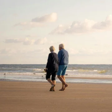 Elderly couple strolling on sandy beach at sunset near seaside hotel with tranquil waves and open sky.