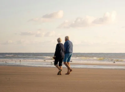 Elderly couple strolling on sandy beach at sunset near seaside hotel with tranquil waves and open sky.