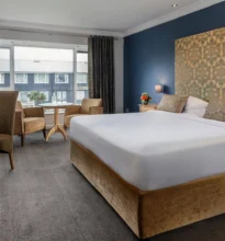 Classic hotel room featuring a large bed with plush white linens, wooden bedside tables, lamps, a cozy chair, and a window with drapes providing natural light.
