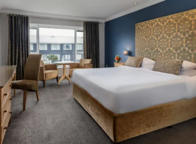 Classic hotel room featuring a large bed with plush white linens, wooden bedside tables, lamps, a cozy chair, and a window with drapes providing natural light.