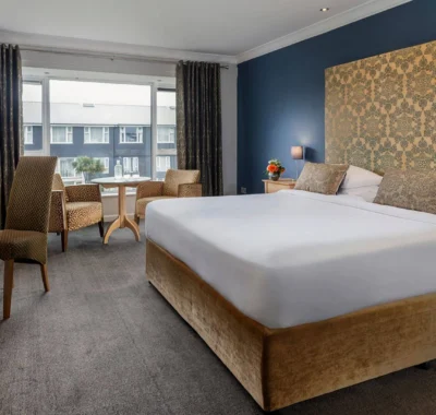 Classic hotel room featuring a large bed with plush white linens, wooden bedside tables, lamps, a cozy chair, and a window with drapes providing natural light.