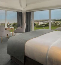Elegant hotel room with a king-sized bed, ocean-view balcony, plush seating area, soft warm lighting, decorated in soothing neutral tones, evoking relaxation and luxury by the coast.