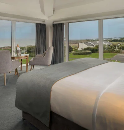 Elegant hotel room with a king-sized bed, ocean-view balcony, plush seating area, soft warm lighting, decorated in soothing neutral tones, evoking relaxation and luxury by the coast.