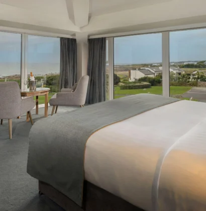 Elegant hotel room with a king-sized bed, ocean-view balcony, plush seating area, soft warm lighting, decorated in soothing neutral tones, evoking relaxation and luxury by the coast.