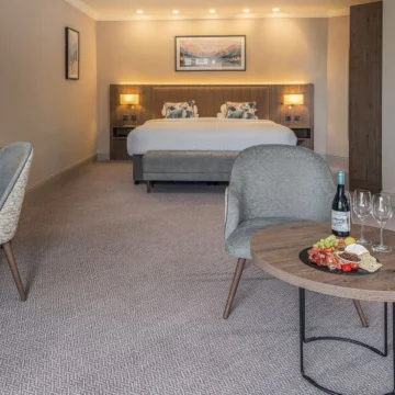 A luxurious hotel suite with a comfortable bed, elegant armchair, and large windows offering a scenic view of the coastal landscape.
