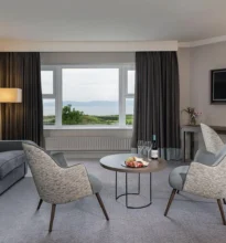 A cozy suite with a large bed, elegant furnishings, a seating area, and a stunning ocean view through a wide window, exuding a serene and luxurious atmosphere.
