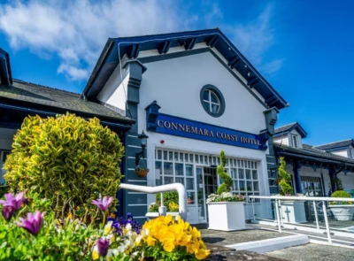 The image shows the entrance of Connemara Coast Hotel, featuring a large glass facade, greenery, and a modern architectural design under a clear blue sky.