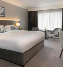 Luxurious hotel room featuring a large double bed with crisp white linens, elegant furnishings, warm lighting, and a scenic view through the window, creating an inviting and cozy atmosphere.